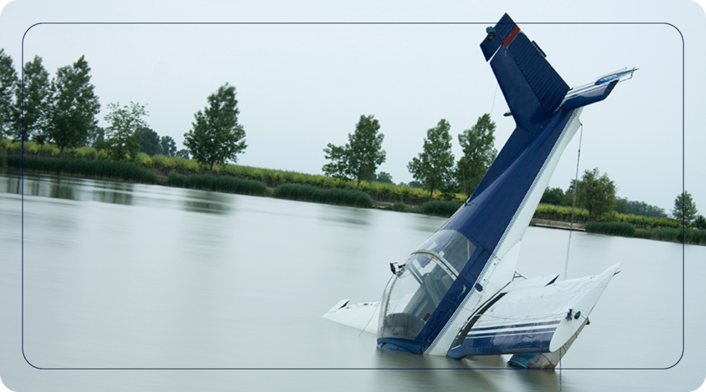 Small aircraft partially submerged nose-down in a body of water with trees in the background.