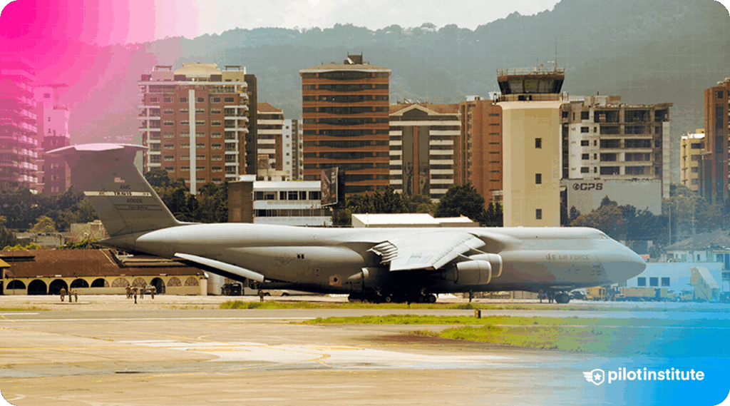 Photo of the Lockheed C-5 Galaxy parked on the tarmac with an airport tower and city buildings in the background.